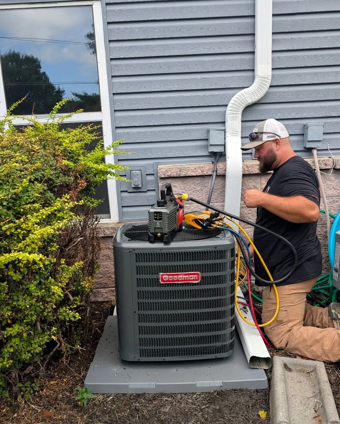 Blake, a Christopherson Mechanical HVAC technician, connecting manifold gauge lines to a Goodman condensing unit alongside a home during an air conditioning service call.